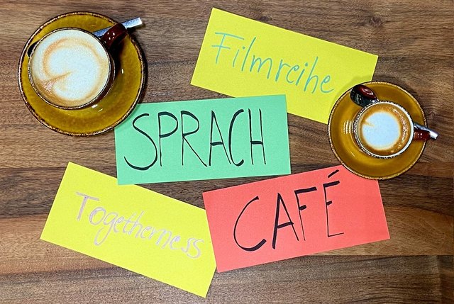 Bild von oben auf einen Tisch fotografiert. Darauf stehen zwei Tassen Kaffee mit Latteart. Auf drei farbigen Zetteln steht: Filmreihe, Sprach, Café, Togetherness